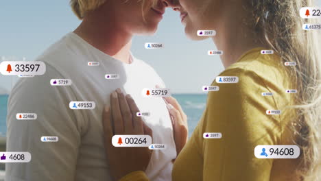 animation of social media icons on banners over caucasian couple in love embracing by seaside