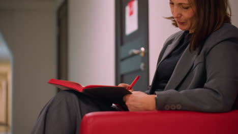 businesswoman writing in notebook, sitting on red armchair in modern office hallway. focused, professional, organizing thoughts, preparing for meeting or brainstorming