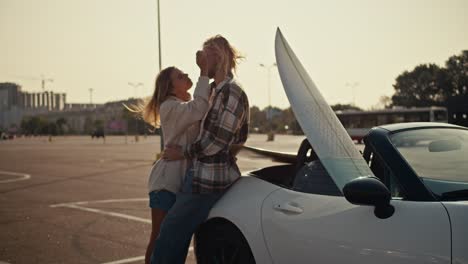 una chica rubia en una sudadera blanca se apoya en su novio rubio en una camisa a cuadros y vaqueros que se sienta en un descapotable blanco y abraza a su novia. pareja feliz, un chico y una chica están de pie cerca de un descapottable blanco y abrazándose en una plaza de la ciudad por la mañana