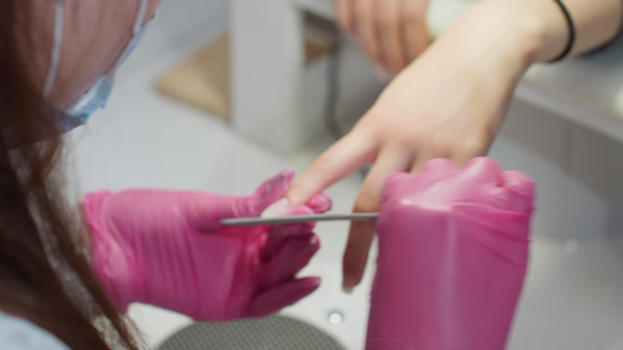 Closeup of nail technician wearing pink gloves carefully filing client fingernails using white nail file, showcasing precision and attention during salon manicure treatment under bright lighting