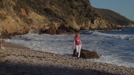 Girl-walking-on-beach,-picks-up-a-stone-from-ground,-strong-waves-crash,-sunny-day