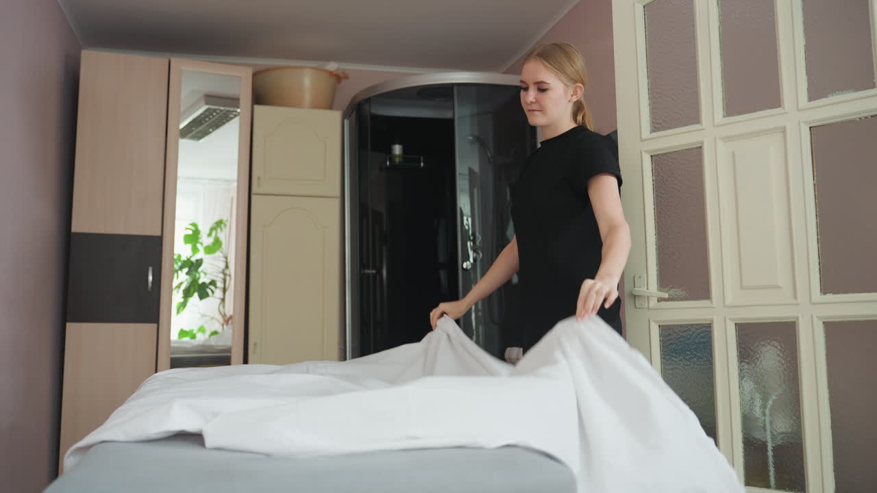 Pretty woman arranges white sheet on bed in well furnished room with glass wardrobe, visible mirror reflection, and soft natural light, showcasing preparation, and calm domestic atmosphere indoors
