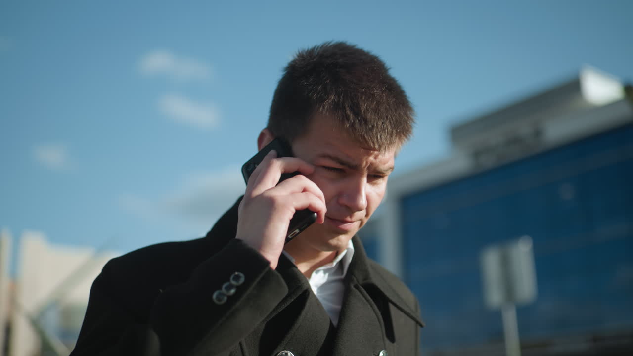 검은 코트를 입은 계약자는 야외에서 전화 대화를 나누고, 의사소통하는 동안 눈에 띄게 머리를 흔들며, 현대적인 유리 건물과 함께 도시 환경에 서 있습니다.