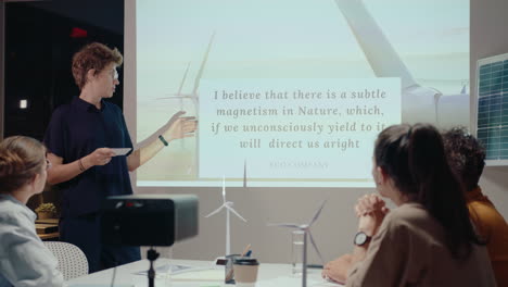 young female engineer standing by projection screen, giving presentation about wind farm to team of colleagues, developing sustainable energy project on night office meeting