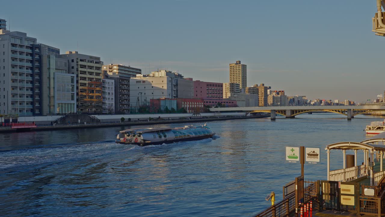 Açık bir günde Tokyo'daki Sumida Nehri boyunca seyir halinde modern fütüristik bir deniz otobüsü. Şehir manzarasını, nehir kenarındaki binaları ve parlak güneş ışığı altında bir köprüyü gösteriyor.