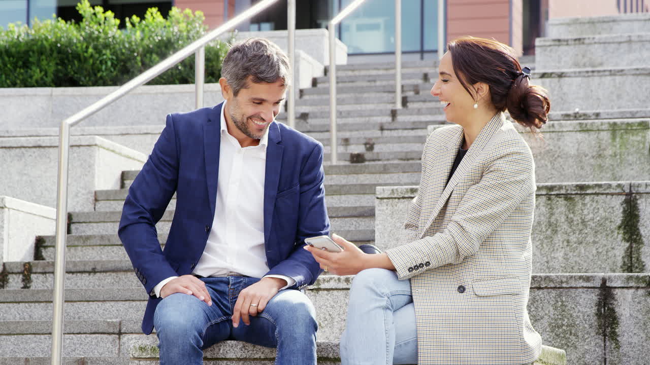 hombre de negocios y mujer de negocios sentados por los escalones teniendo una reunión al aire libre mirando el teléfono