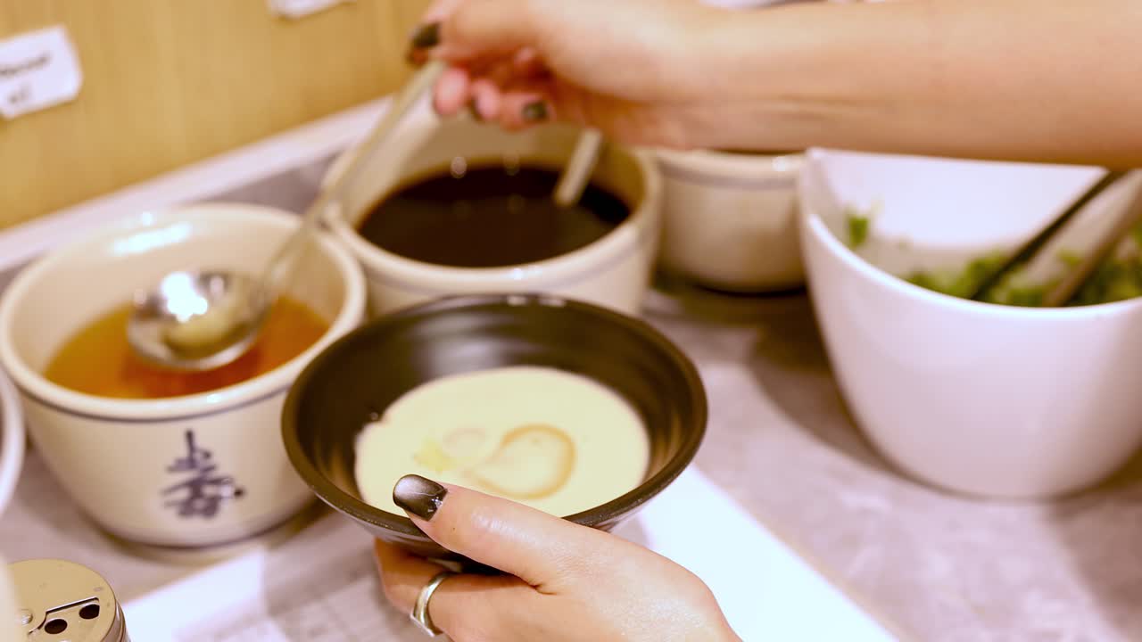 Person ladles spicy sauce into bowl at self-service condiment station, warm indoor lighting, close-up