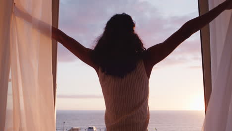 mujer feliz abriendo la cortina disfrutando del amanecer temprano en casa sintiendo una libertad pacífica relajándose en vacaciones mirando la hermosa vista del océano