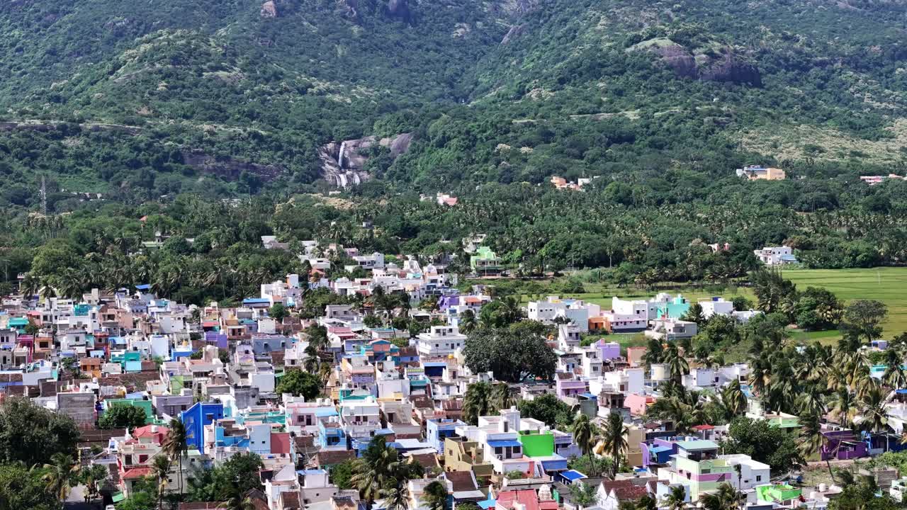 A majestic famous Courtallam (Kutralam) Main Falls in Tamil Nadu, India. cascades down a rocky, forested mountainside. A settlement is nestled below in the lush valley
