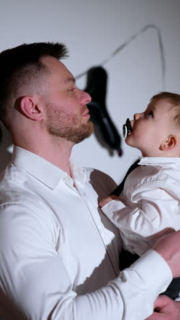 man in white shirt holding a little baby boy in white shirt and black tie. blond woman in black dress comes up holding balloons. vertical video.