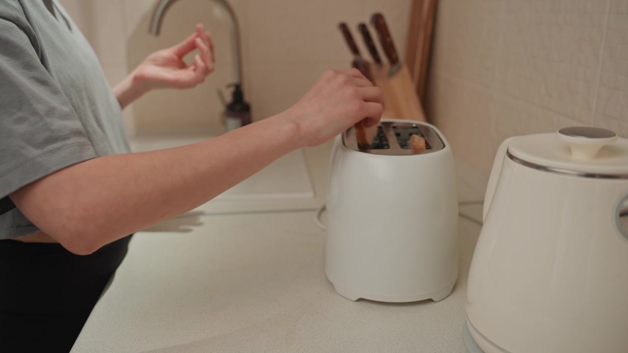 Person placing toast into toaster morning kitchen, cropped torso and hands, casual shirt, kettle and utensils on counter, upbeat rhythm vibe, music playing, playful domestic moment, quick breakfast