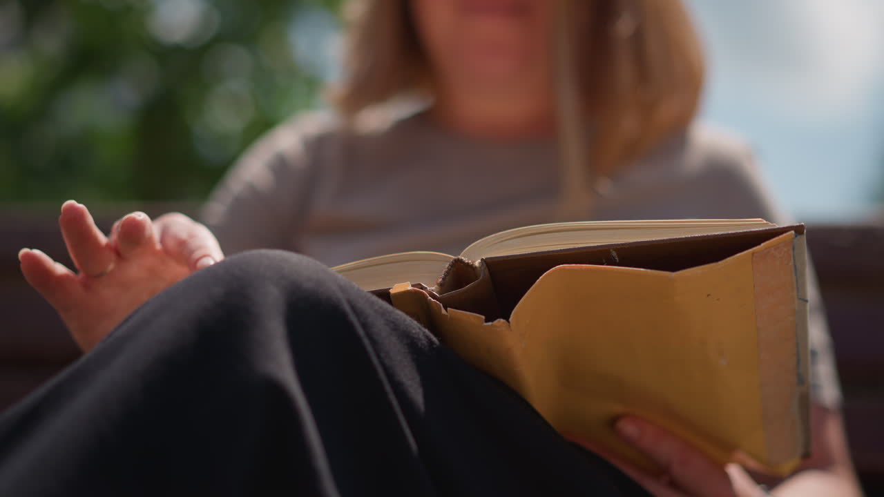 Close up of woman sitting outdoors flipping to new page in worn book under bright blue sky with blurred greenery in background calm sunlight peaceful summer moment showing relaxation