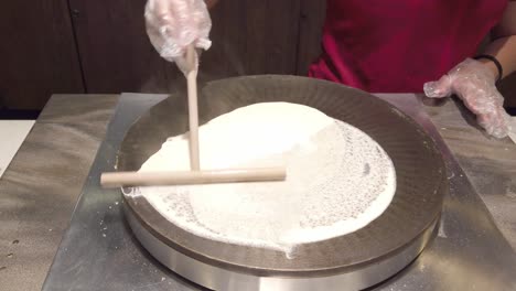 gloved hands evenly spread crepe batter in a circular motion on a round griddle using a wooden spreader