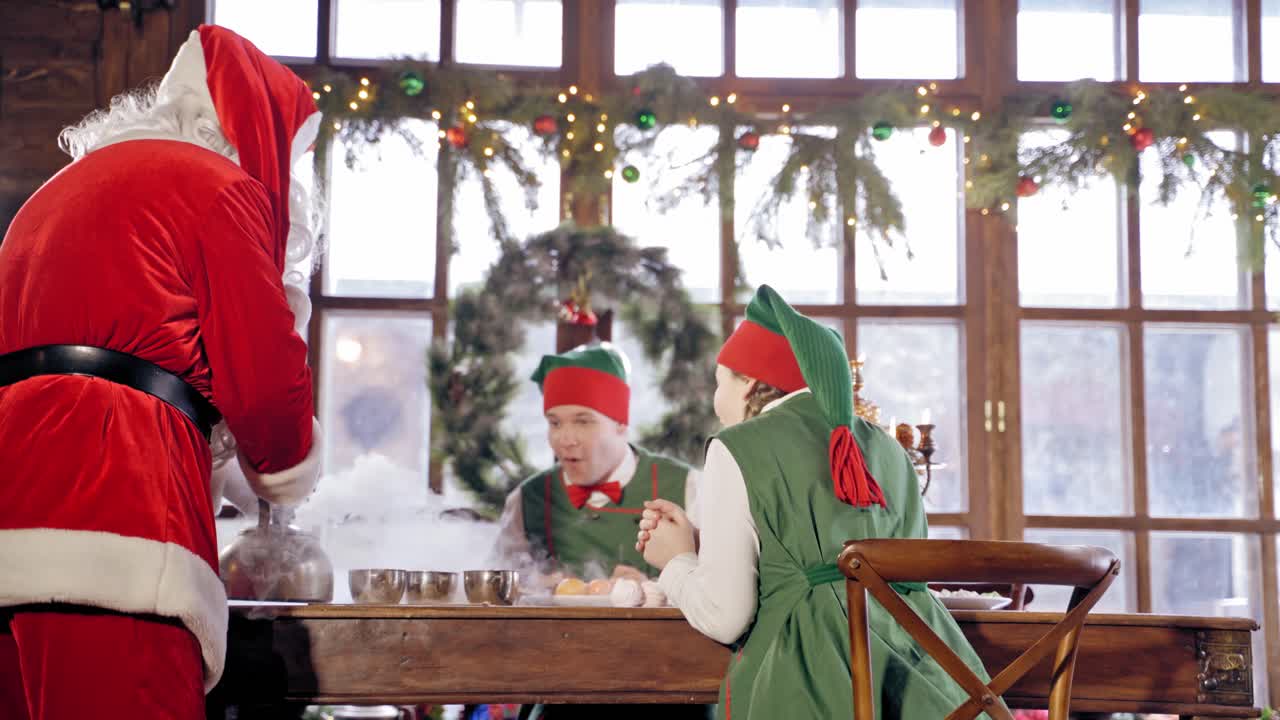 Santa Claus is pouring hot tea to the cups in specially decorated room. Happy elves are looking at the process. New Year and Christmas concept. Feast is coming.