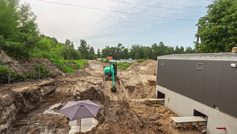 -High-angle-shot-of-green-excavator-leveling-earth-for-construction-of-a-parking-lot-beside-newly-made-building-throughout-the-day-in-timelapse