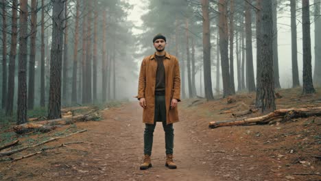 young male hiker wearing brown coat and beanie is standing on a path in a misty forest during a cold autumn day, surrounded by tall pine trees and fallen branches