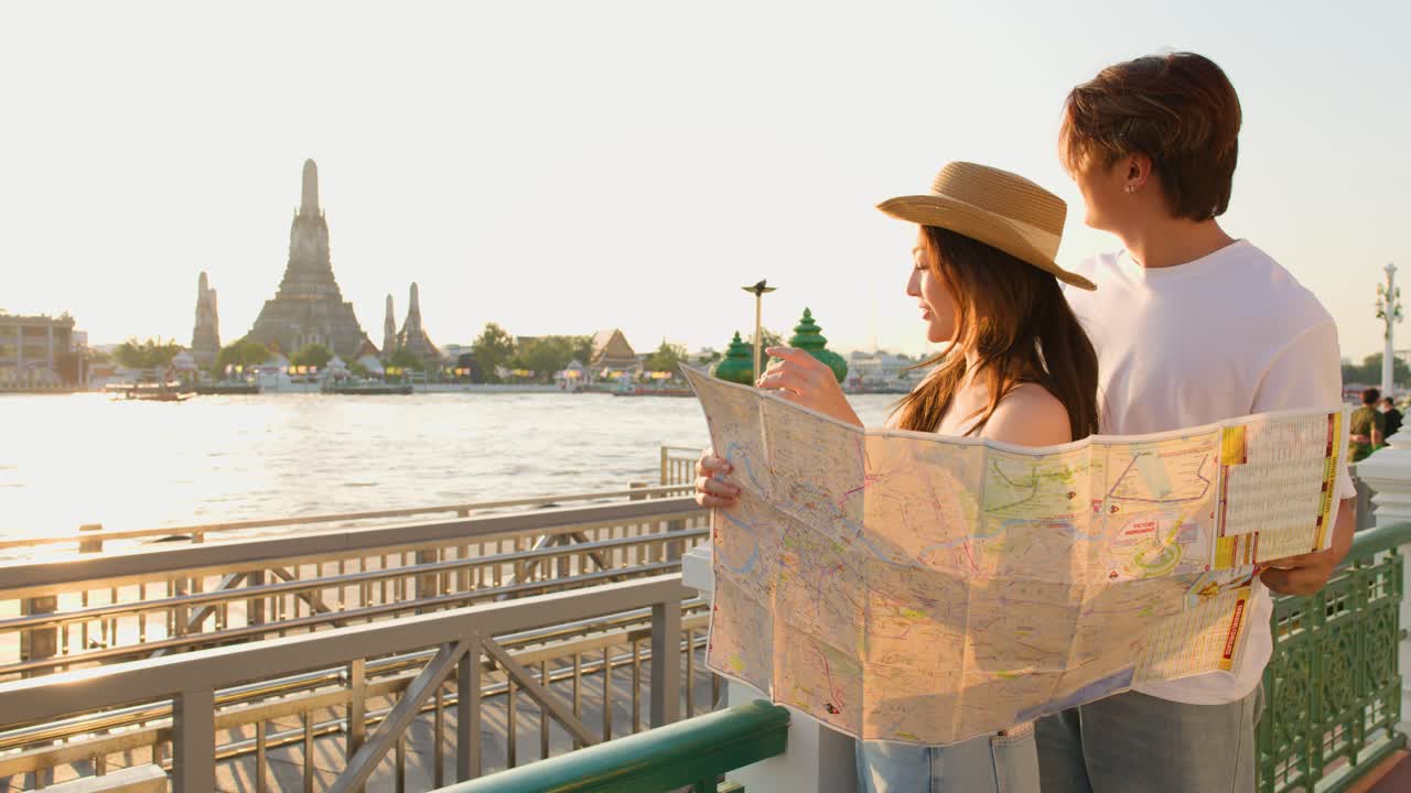 Coppia asiatica consulta mappa vicino al fiume, tempio Wat Arun visibile, illuminazione calda del tramonto, atmosfera rilassata.