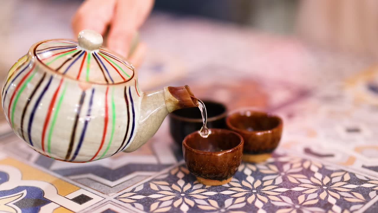 A hand pours tea from a striped teapot into small cups on a patterned tablecloth, creating a warm, inviting atmosphere