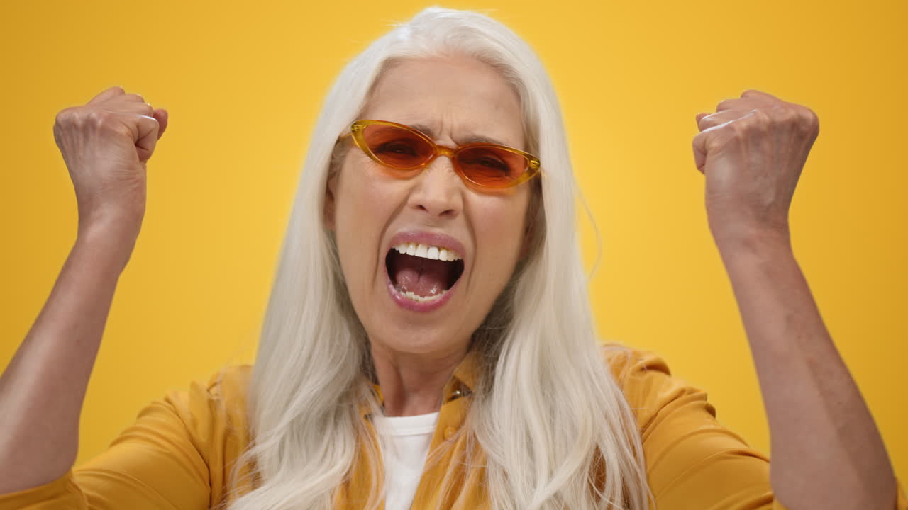 Furious senior woman shouting in studio. Angry old lady raising fists inside.