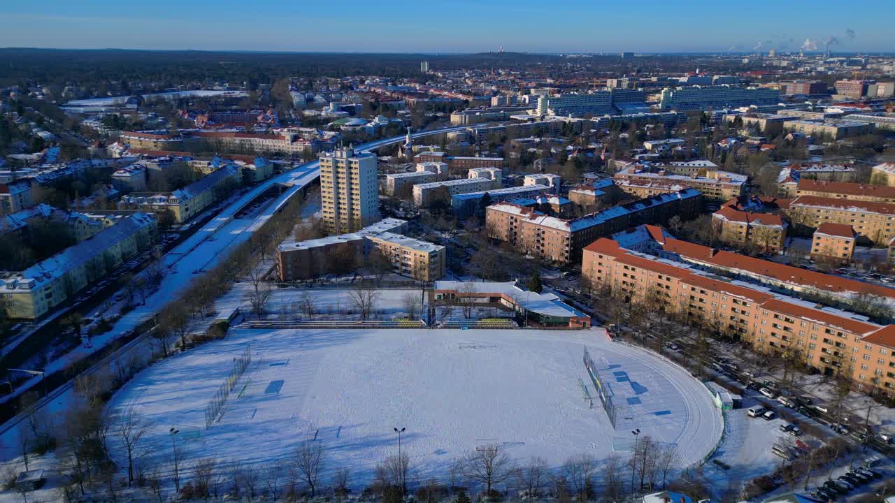 Saksan pääkaupungin maisema, jossa on lumen peittämä jalkapallokenttä ja juoksurata asuinrakennusten ympäröimänä talvella. Upea panoraamanäkymä ilmasta, staattinen kolmijalka leijuvan dronen avulla