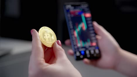 a woman examines the physical golden bitcoin coin while checking the cryptocurrency trading app on her smartphone