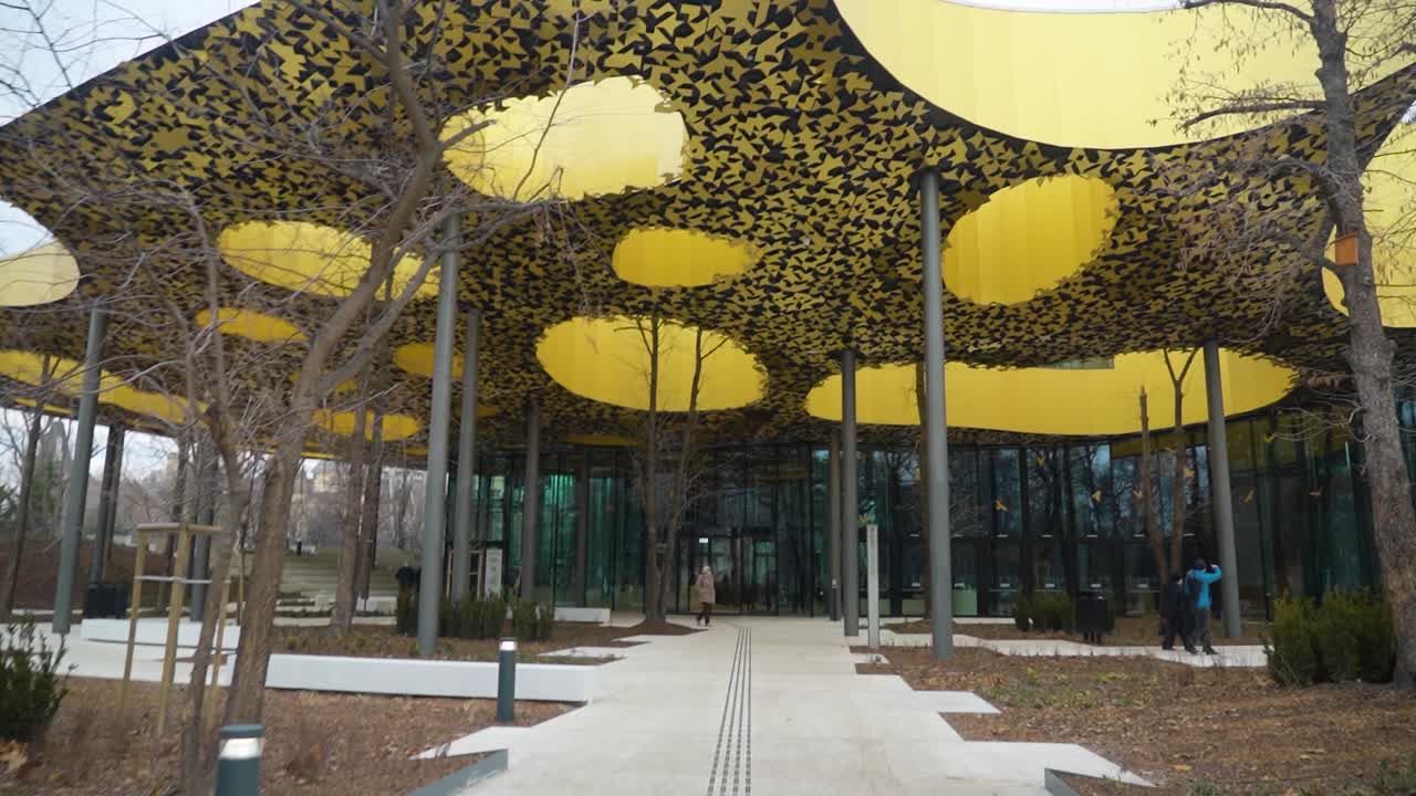 современный дом музыки - венгерское здание с уникальной золотой крышей и стеклянными стенами, окруженное деревьями в ландшафтном саду.