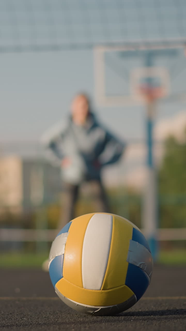 노란색 표시가 눈에 보이는 야외 스포츠 경기장에서 바닥에 놓인 배구, 흐릿한 배경의 운동 선수들이 배구와 운동에 초점을 맞추고 체력 운동에 참여합니다.