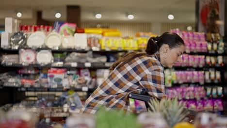 a brunette girl in a plaid shirt puts fruits in a cart during her shopping in a supermarket. confident brunette woman chooses oranges during her shopping