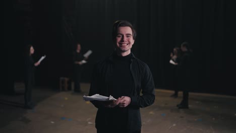 Portrait-of-a-happy-young-male-actor-in-a-black-suit-with-papers-in-his-hands-against-of-actors-who-are-rehearsing-for-a-performance-on-stage-in-a-theater-with-black-curtains