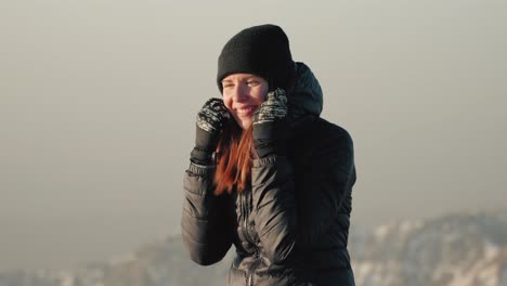 young caucasian woman in beanie adjusting her winter jacket during sunrise on top of a mountain