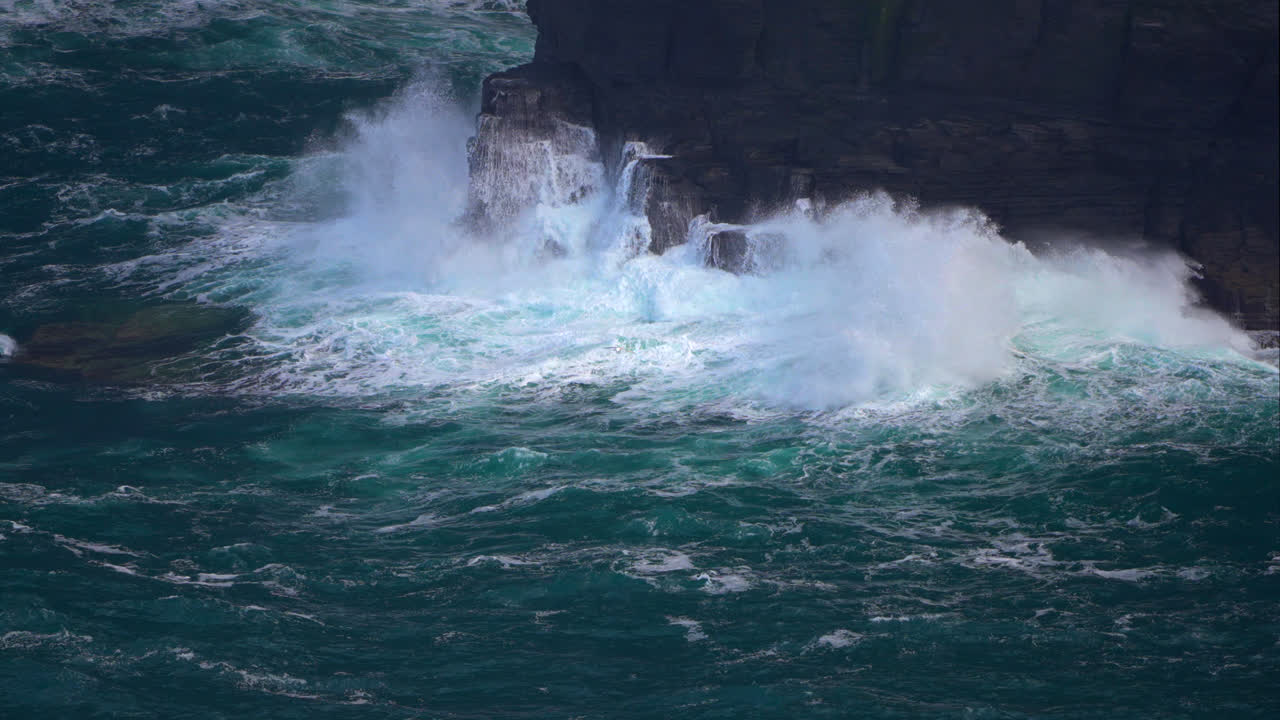 Powerful ocean waves crash against the base of the Cliffs of Moher, sending white spray into the air. The rugged coastline and towering cliffs showcase the untamed beauty of Ireland’s west coast.