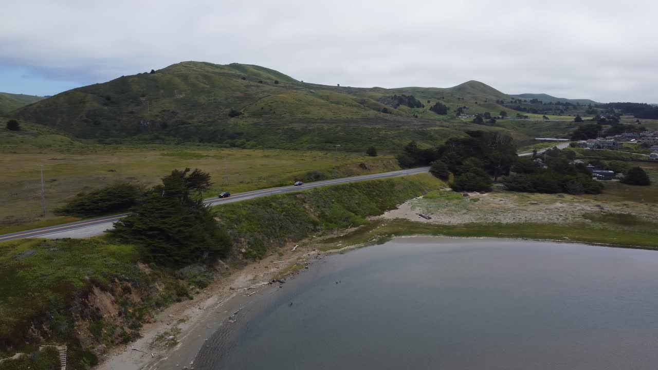 Cars driving up the coastal highway in Northern California as rolling green hills dotted in trees fill the background. Overcast drone shot near a town and the water in 4K.
