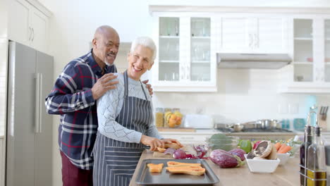 happy senior diverse couple preparing vegetables and embracing in kitchen, copy space, slow motion