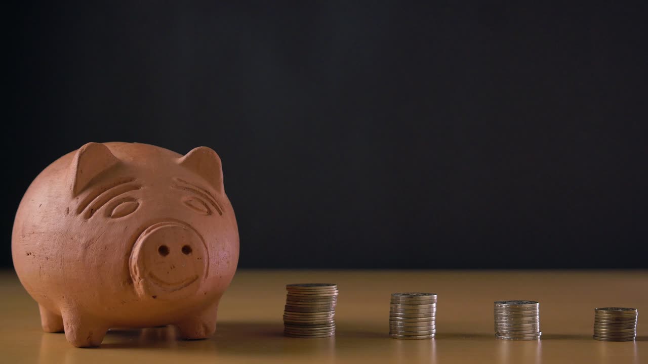 Hand of a white man putting coin in a clay piggy bank and stacks of coins