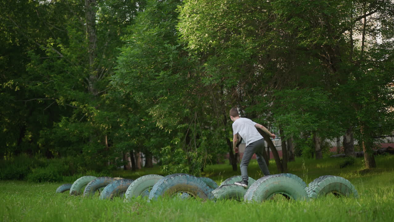 백색 셔츠와 회색 바지를 입은 어린 소년이 잔디에 배치된 타이어를 가로질러 뛰어다니며, 배경에는 울창한 나무가 둘러싸여 있습니다.