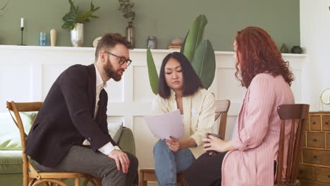 Young-Woman-Holding-Paper-Report-And-Debating-With-Her-Two-Colleagues-While-Sitting-In-Wooden-Chairs-In-A-Living-Room