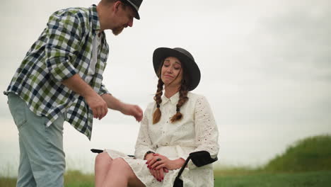 in a serene grassy field, a craftsperson in a checked shirt adjusts the hair of a woman seated in a chair, wearing a white dress and black hat