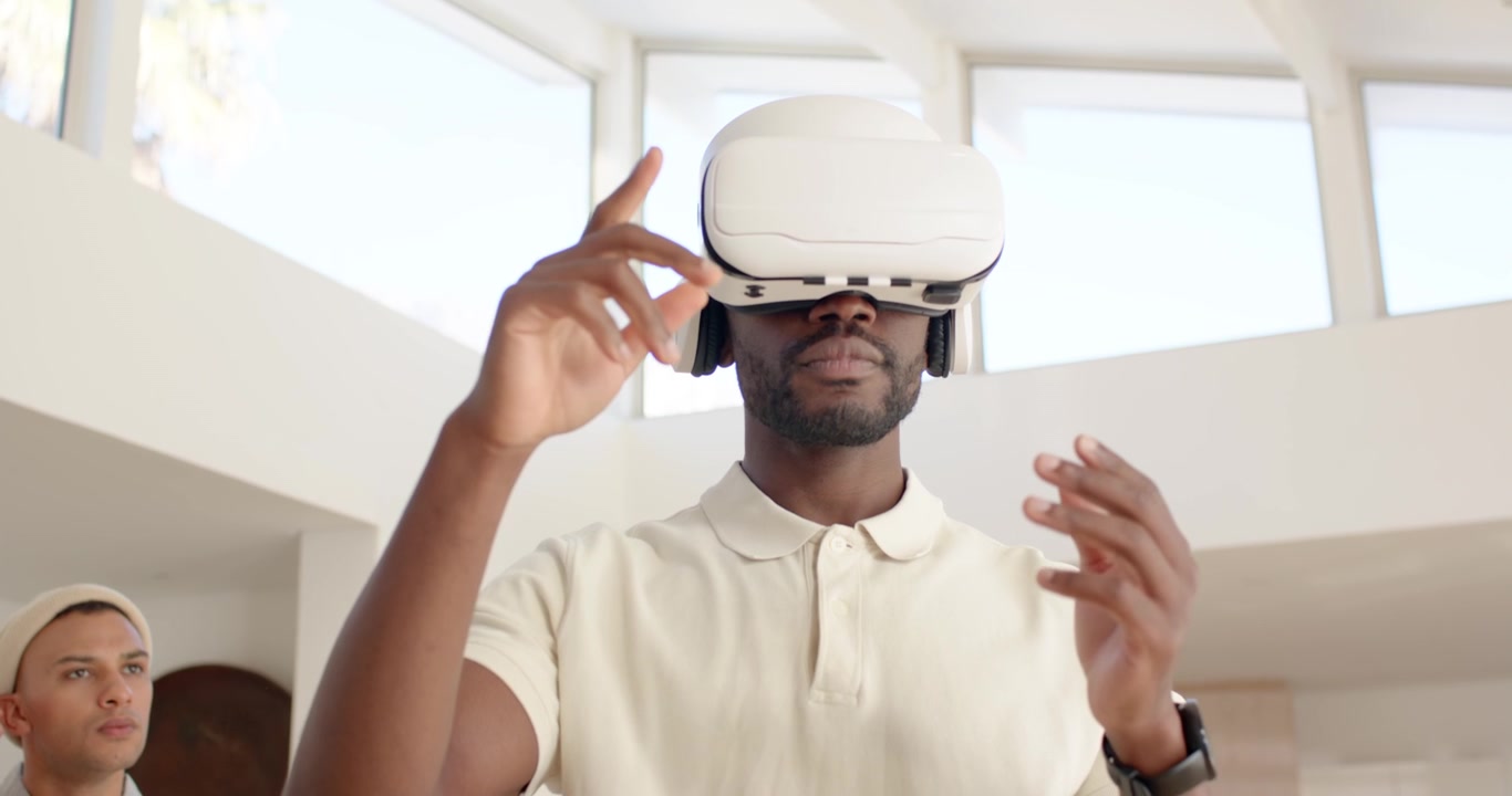 Uomini di diverse etnie che guardano un uomo sollevare un visore VR bianco, testando una scena mentre un uomo digita al laptop in cucina.