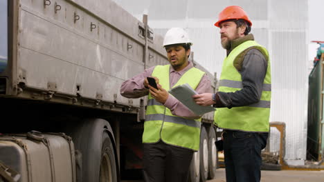 Chef-Und-Arbeiter-Mit-Westen-Und-Schutzhelmen-Organisieren-Eine-Lkw-flotte-In-Einem-Logistikpark,-Während-Sie-Auf-Einem-Smartphone-Lesen-Und-Dokumente-Lesen