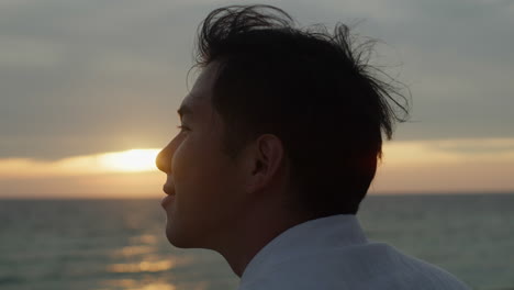 젊은 아시아 남성이 바다 앞에서 해가 지는 것을 즐기고 있습니다. 바다, 여름의 진동, 부드러운 바람, 근접 초상화 모델.