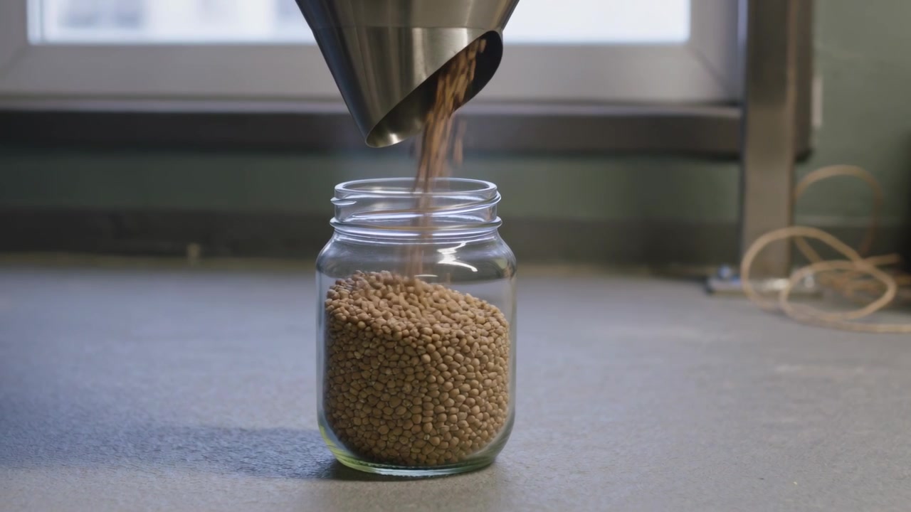 Riempimento di un barattolo di vetro trasparente che si alza sul bancone, con un ugello d'acciaio che si apre e eroga pellet beige.