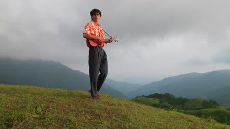 Low-Angle-Aufnahme-Eines-Asiatischen-Mannes,-Der-An-Einem-Regnerischen-Tag-Auf-Einem-Berg-Läuft-Und-Dabei-Ukulele-Spielt