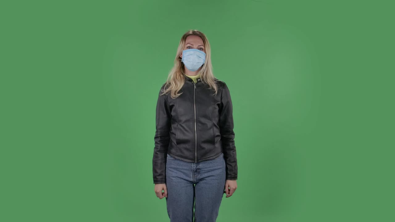 Portrait of beautiful young woman in medical protective face mask looking at camera and pointing fingers to the side. Blonde with loose hair in a black jacket and jeans on a green screen in the studio. Health Protection Corona Virus Concept