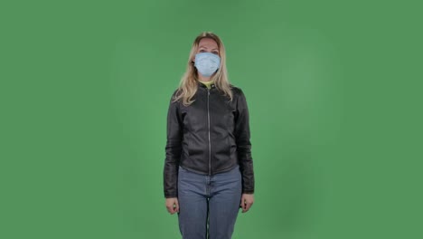 portrait of beautiful young woman in medical protective face mask looking at camera and pointing fingers to the side. blonde with loose hair in a black jacket and jeans on a green screen in the studio. health protection corona virus concept