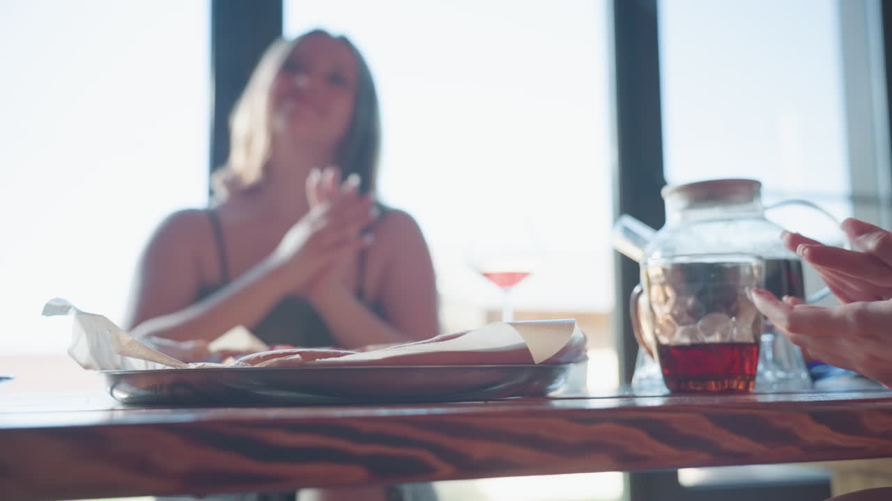 Friends having good time, one woman drops pizza on table while smiling, holding pizza in hand, drink in glass kettle nearby, towel tied around her waist, cozy indoor setting with wooden walls