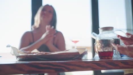 friends having good time, one woman drops pizza on table while smiling, holding pizza in hand, drink in glass kettle nearby, towel tied around her waist, cozy indoor setting with wooden walls