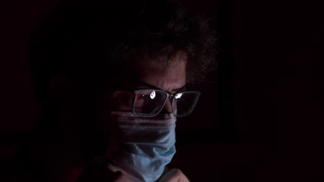 Young-bearded-male-wearing-glasses-adjusting-surgical-corona-virus-mask-in-darkness