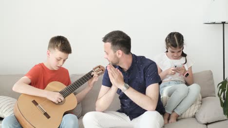 a child plays the guitar for his father and sister. a boy demonstrates playing an acoustic guitar. family relaxing at home on sofa