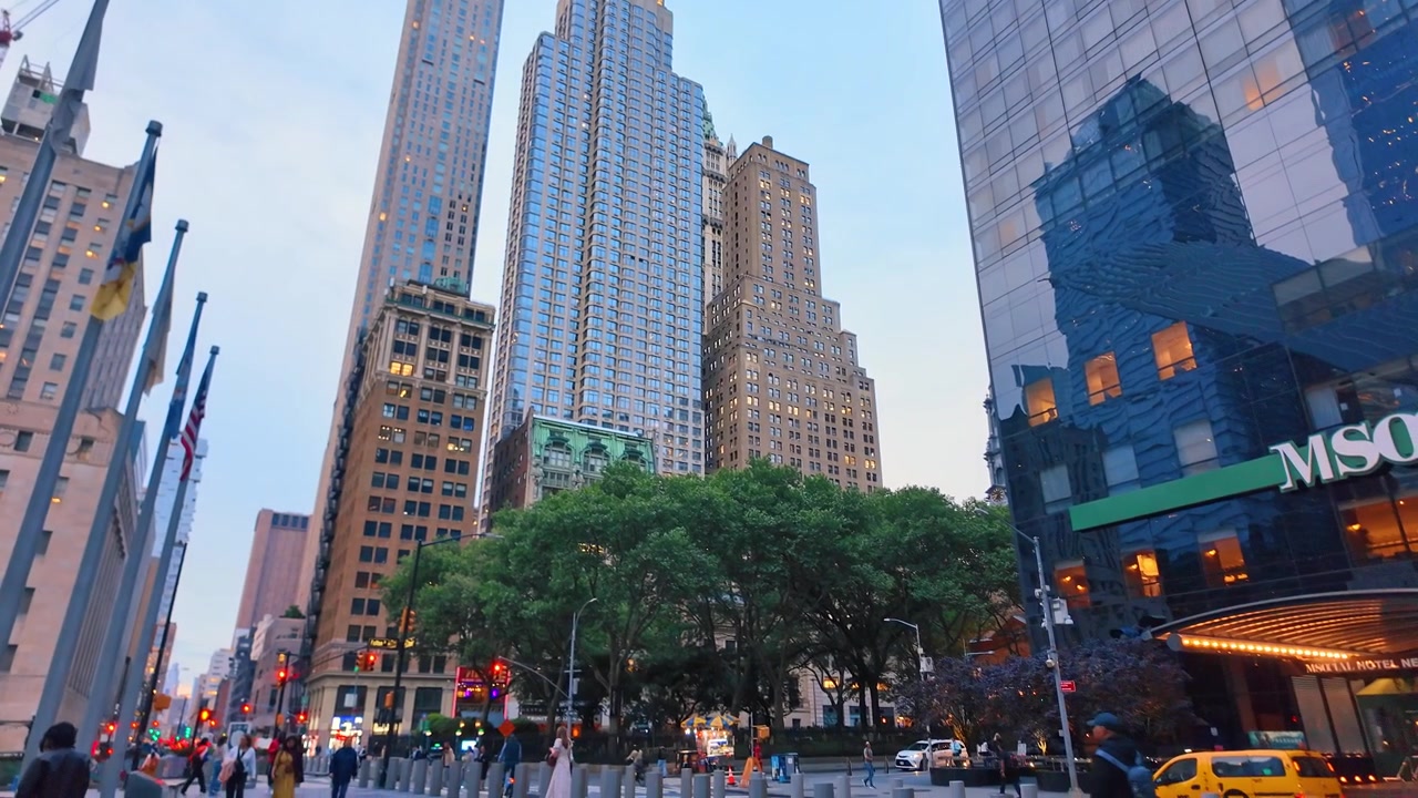 New York, USA, 5 giugno 2026: Grattacieli di epoche diverse fiancheggiano Broadway a Lower Manhattan. La vista stradale contrappone torri di vetro a facciate in pietra calcarea e una chioma di alberi sotto un cielo crepuscolare pallido.