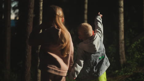 children look at wild forest at evening twilight. boy points forward paying sister attention to nature in night woods. little kids at woodland resort